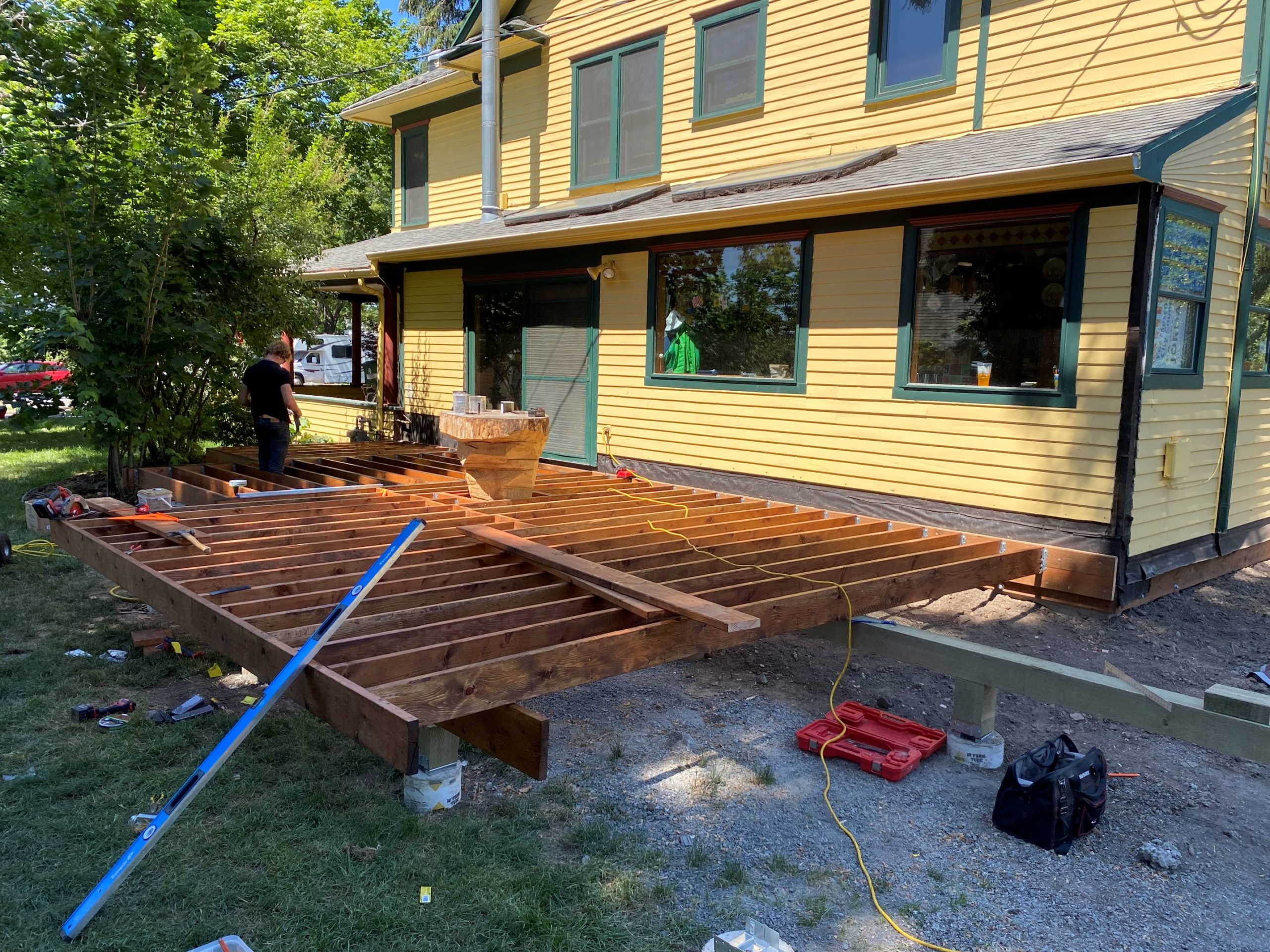 Deck framing under construction alongside a yellow home